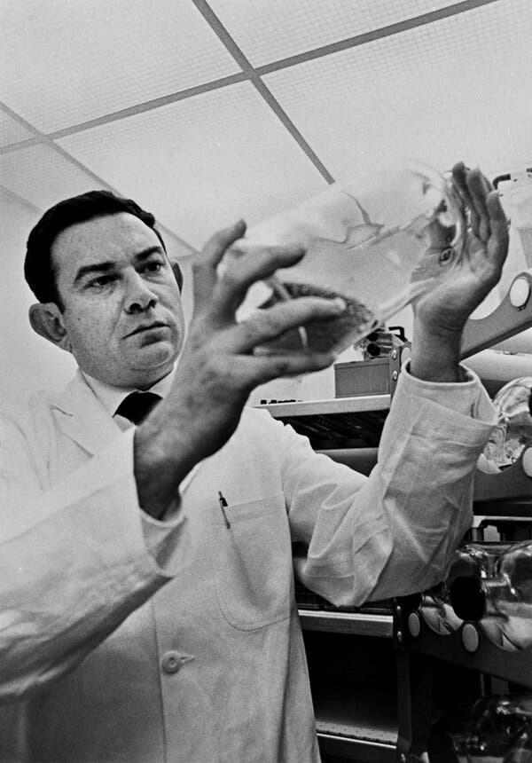 A black-and-white photo of Leonard Hayflick, a man in a white lab coat, holding up a glass container and looking at it intensely.