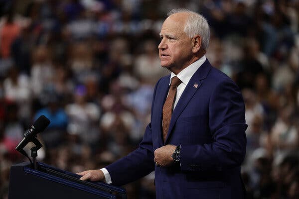 Tim Walz stands at a lectern, wearing a blue suit and a tie.