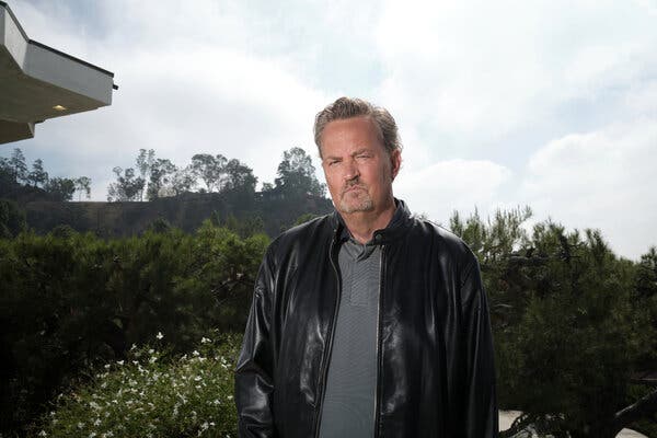 Matthew Perry, with a mustache and goatee, stands outdoors in front of some trees in a black leather jacket and a gray shirt.