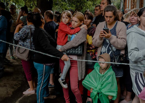 A crowd of people, including several women holding children, behind a rope line.