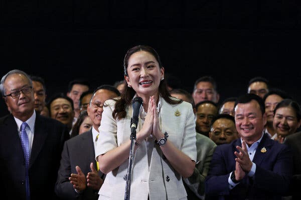 A woman in a white suit, flanked by smiling men, stands at a microphone.