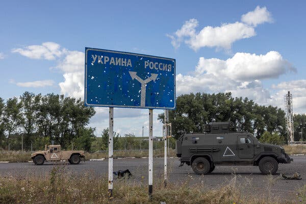 A sign pockmarked with bullet holes directs drivers toward Ukraine and Russia. Two military vehicles are on the road.