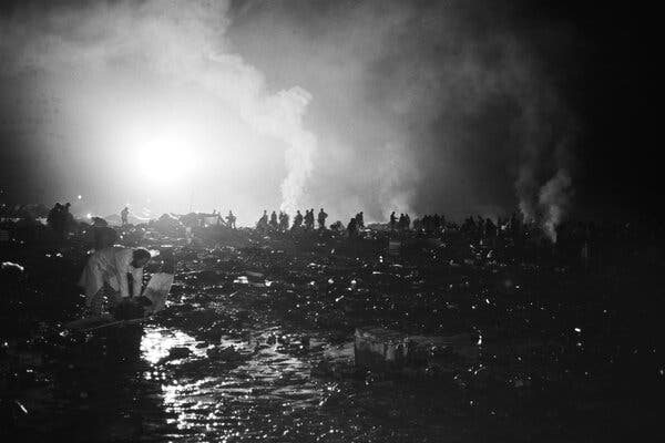 Smoke rises from a field where people, silhouetted by light, walk about. 