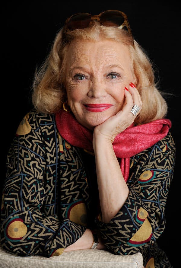 A close-up portrait of Gena Rowlands, who is gazing directly at the camera. She has blond hair and sunglasses perched on her head. She is wearing a patterned blouse and a pink scarf and is resting her chin on her left hand.