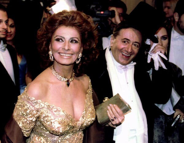 Sophia Loren, in a gold strapless dress, holds a handbag while smiling next to a man in a tuxedo.