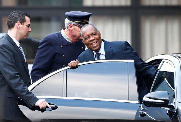 Issa Hayatou, formally dressed, gets into a car as one man holds the door open for him and another, in uniform, stands behind him.