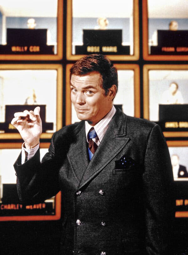 In a close-up promotional photo, he looks at the camera while pointing his thumb over his shoulder at celebrity guests seated in a grid of squares. He has on a gray double-breasted suit jacket with wide lapels.