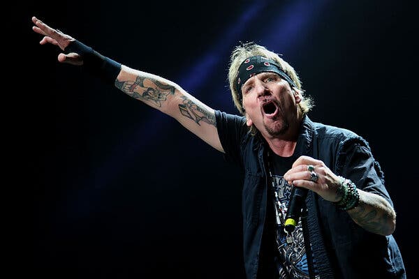 A man holds a microphone while onstage, wearing a bandanna. His arms are tattooed.