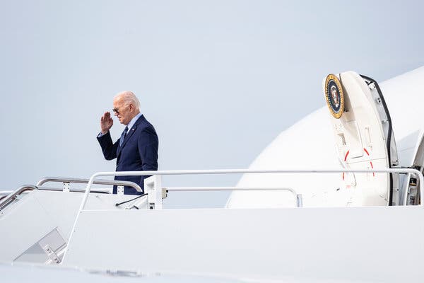 President Biden, wearing a blue suit and tie and aviator sunglasses, standing on the stairs of Air Force One. His hand is raised as if in a salute. 