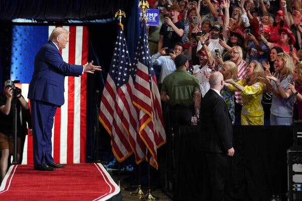 Donald J. Trump on a stage gesturing at a crowd of supporters in front of him.