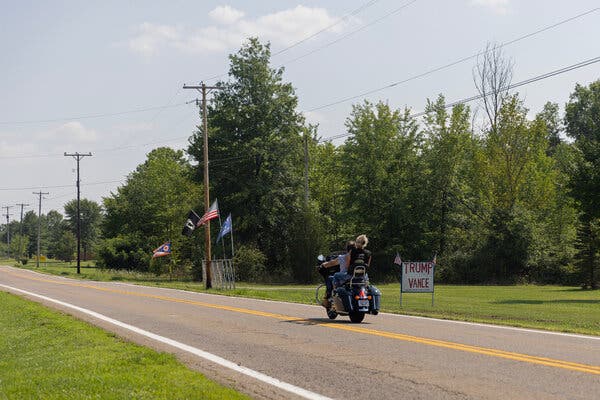 A couple ride a motorcycle along a rural road past a sign that reads, ‘Trump Vance.’