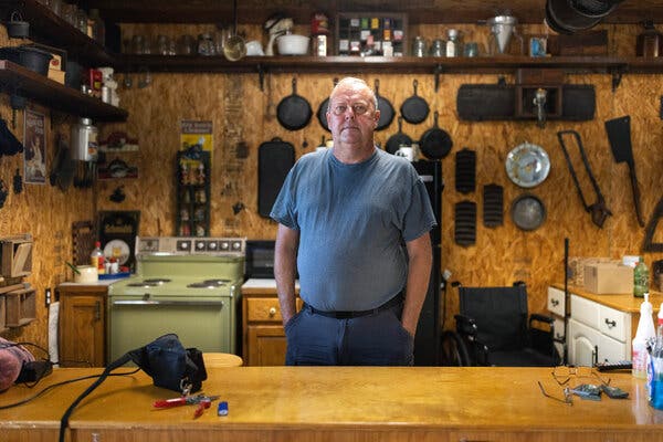 A man standing behind a counter with a stove in the background and pans hanging on the wall behind him.