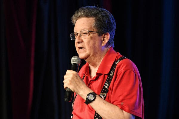 Perry Kurtz speaks into a wireless microphone while performing onstage during a comedy routine. He is wearing a red polo shirt with suspenders that feature a peace sign pattern on them.