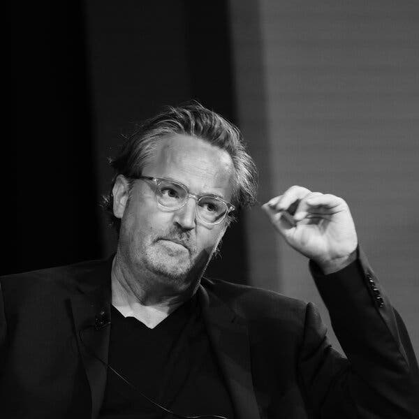 A black-and-white photograph of Matthew Perry tilting his head and reaching for his eyeglasses. 