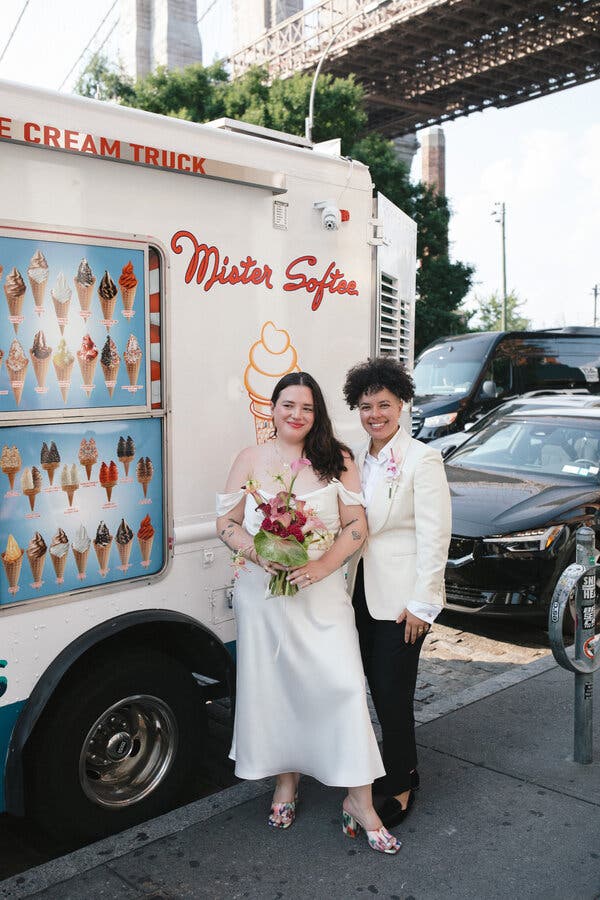 Two women stand in front of a Mister Softee truck in wedding attire. The woman, left, wears an-off the-shoulder white ankle-length gown and is holding a bouquet of red and pink flowers. The other woman, beside her, wears a white jacket, with a flower in her lapel, over an open collar white shirt and black pants. 