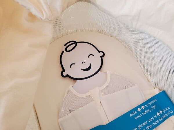 A cardboard cutout of a baby is strapped into a new Snoo smart bassinet that is still outfitted with safety labels.