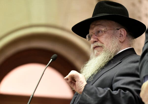 Rabbi Shmuel Butman gestures with his left hand as he speaks to New York State lawmakers in 2017. He has a long gray beard and wears a black hat, eyeglasses and a black suit.