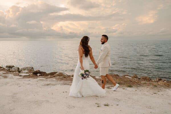 A couple with their backs to the camera hold hands and look at each other while walking along a sandy beach under a cloudy sky. The bride, left, in a white gown, holds a bouquet of white and lilac-colored roses. The groom wears an ivory-colored jacket with matching shorts and white sneakers. He has a tattoo on the side of one leg. 