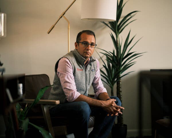 Sean Ammirati, wearing a gray fleece vest, sits in a chair indoors, facing the camera, his hands clasped in front of him. Two potted palms are behind him.