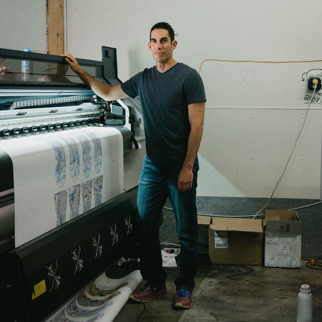 Steven Bright, facing the camera, has his right hand on the top of a dye sublimation printer from which a roll of paper is emerging with mitten designs.