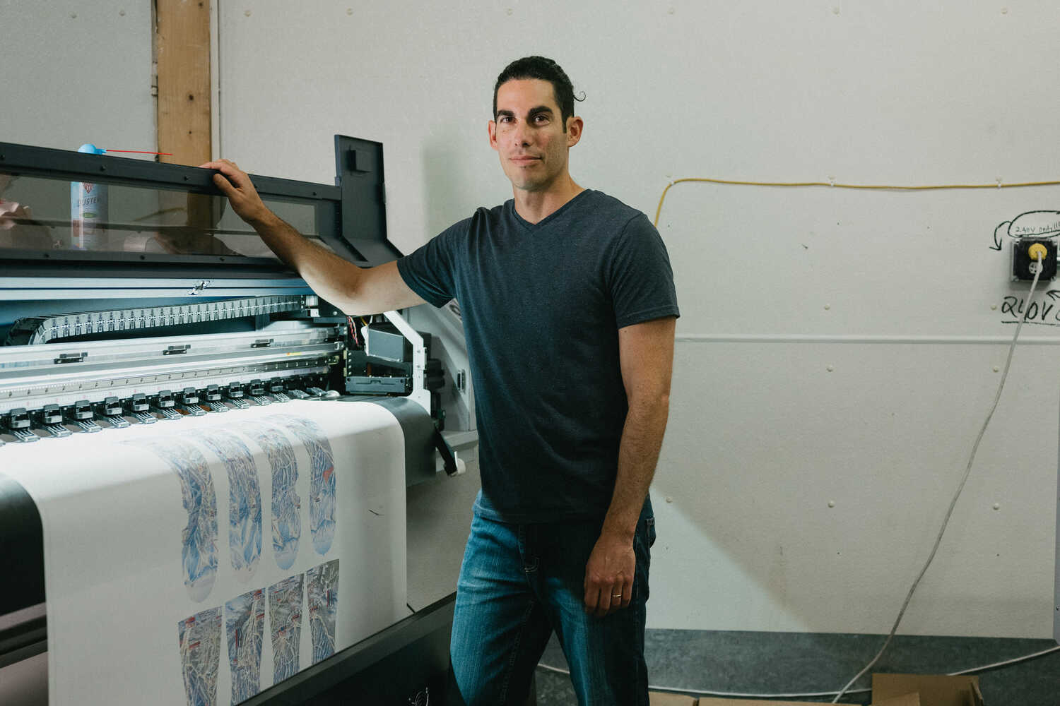 Steven Bright, facing the camera, has his right hand on the top of a dye sublimation printer from which a roll of paper is emerging with mitten designs.