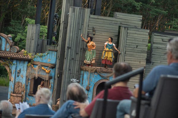 Audience members look on as two actresses wearing dresses stand on a second-level balcony onstage. Another actress can be seen standing in a window in the lower left of the stage.