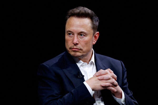 Elon Musk, wearing a suit with no tie, sits onstage with his fingers interlaced.