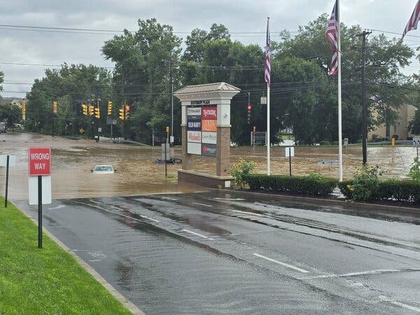 A shopping center sign next to a flooded intersection where several cars are nearly submerged in brown water.