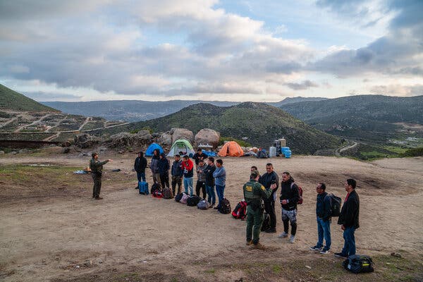 Two people in green uniforms speak to a group of men who are lined up overlooking a hilly landscape.