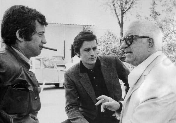 In a black and white photo, three men stand talking to each other. 