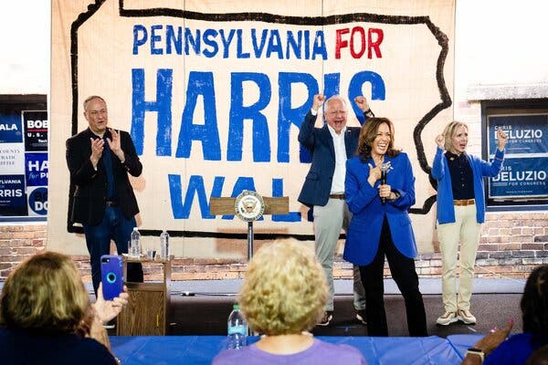 Kamala Harris stands on stage with Tim Walz and their spouses. There is a sign behind them that reads Pennsylvania for Harris Walz.