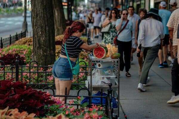 A woman with red hair dressed in a black and white top and jean shorts sells fruit from a cart on a street filled with pedestrians. 