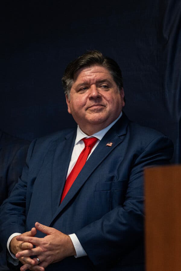 JB Pritzker, in a blue suit and a red tie, smiling and interlacing his fingers.