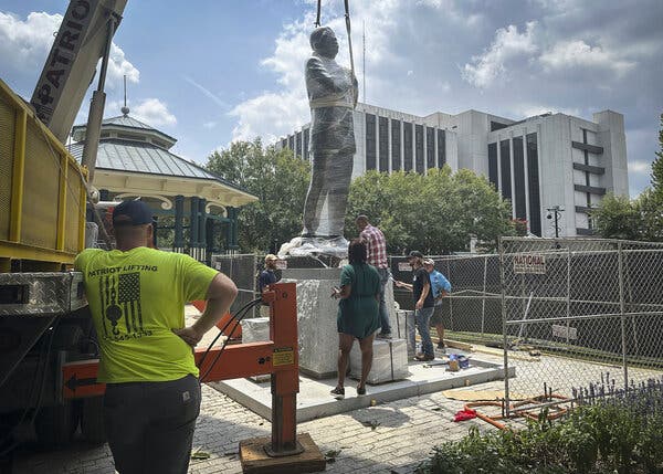 A statue of John Lewis, which is wrapped in clear plastic, is lowered into place by a crane. Workers stand around the base of the statue.