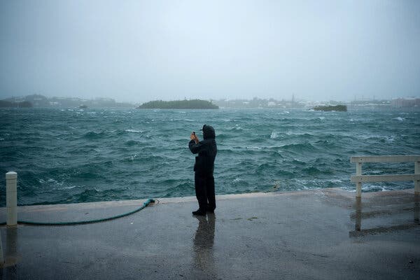 A lone figure in rain gear stands at the edge of a harbor with turbulent blue waters.