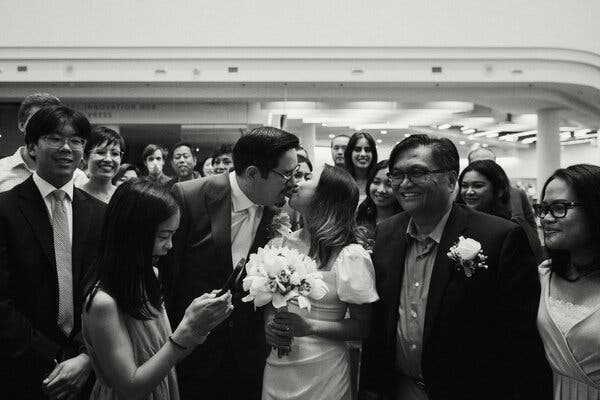 The couple are kissing, and surrounded by smiling friends and family at the Toronto Reference Library in a black and white photo. The groom, left, is wearing a suit and tie, while the bride, wearing a gown with short puffy sleeves, is holding a bouquet of orchids. 