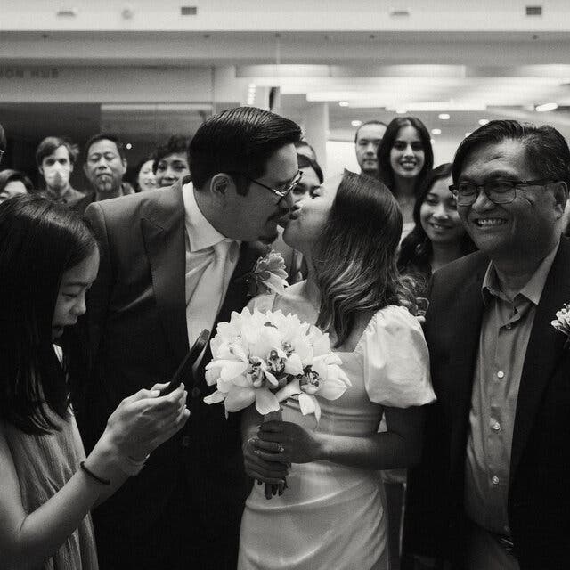 The couple are kissing, and surrounded by smiling friends and family at the Toronto Reference Library in a black and white photo. The groom, left, is wearing a suit and tie, while the bride, wearing a gown with short puffy sleeves, is holding a bouquet of orchids. 