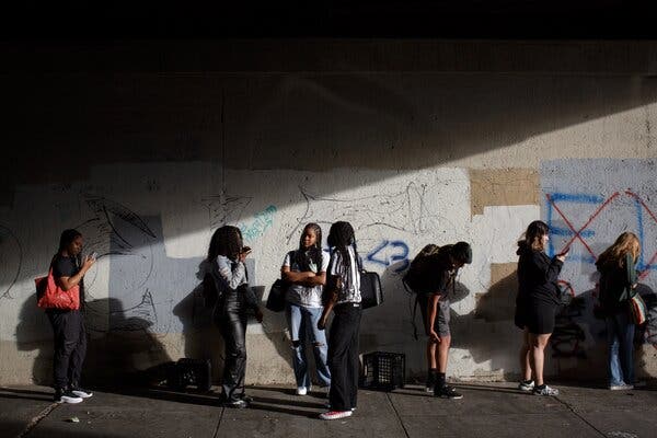 Urban high-school students waiting against a heavily-graffitied wall. A group of three girls face on another in conversation; the other students stare at their phones.