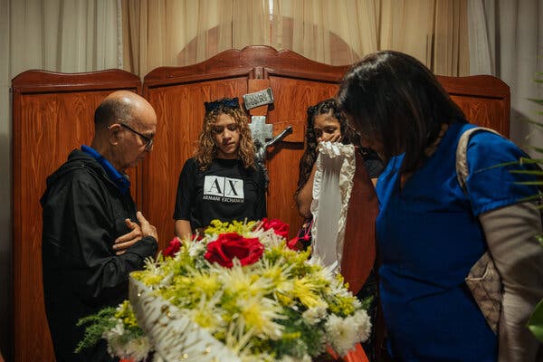 Three women and a man stand looking at a coffin draped in yellow and red flowers.