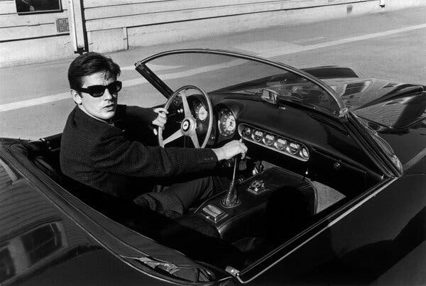 A black and white photo of Alain Delon driving a convertible sports car in sunglasses, looking at the camera with one hand on the steering wheel and the other on the gearshift.