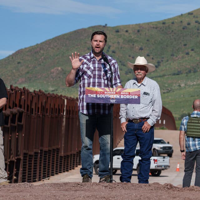 JD Vance speaking at a lectern along the border. Two men are behind him watching him speak, and a third man looks away into the distance.