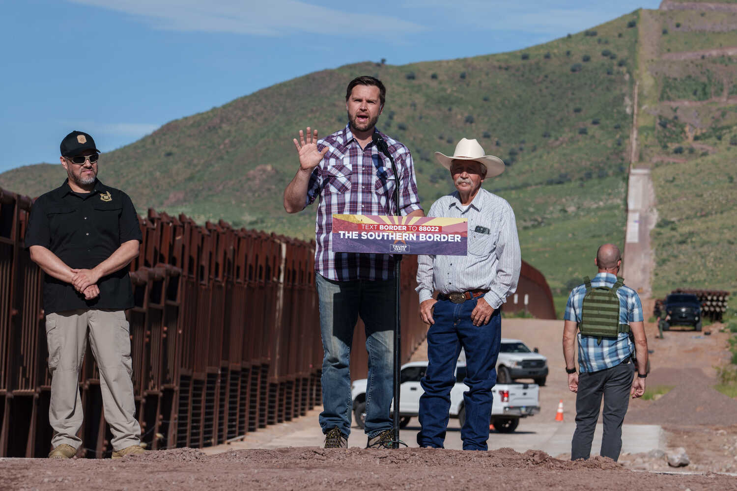 JD Vance speaking at a lectern along the border. Two men are behind him watching him speak, and a third man looks away into the distance.