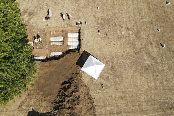 An aerial view of a cemetery with a big hole in the dirt and some white, rectangular caskets in the ground. Workers wearing reflective gear and a white tent are next to the hole.