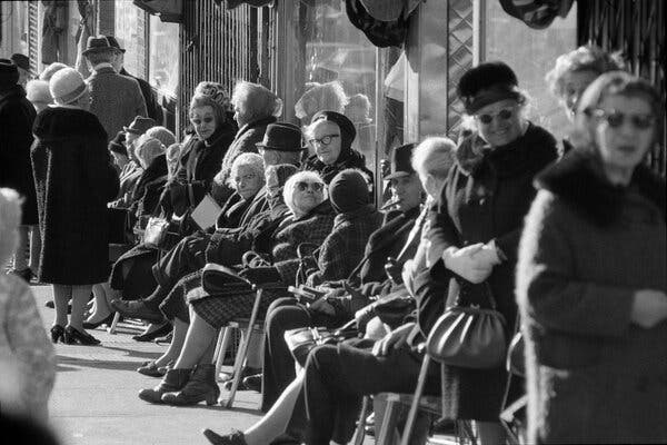 A black-and-white photograph from 1970 shows many elderly white women, and a few elderly men, dressed for winter while sitting and chatting in front of stores on a sunny Bronx street.