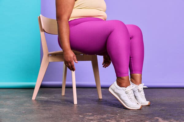 A woman in pink workout pants and white sneakers sits in a wooden chair sits on a workout mat in front of a blue and purple backdrop.