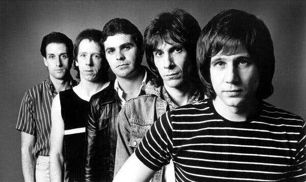 A black-and-white publicity photo of five musicians, all standing and all with expressionless faces. Greg Kihn, wearing a striped T-shirt, is on the right.