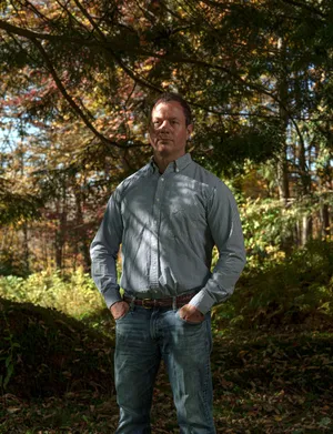 Renny Babiarz, wearing a button-down shirt and jeans, stands with his hands in his pockets in the woods.