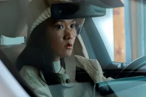 Ali Wong in the driver’s seat of a car. She wears a tan sweater, a cream knit bucket hat and large wire-rim glasses.