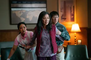 Michelle Yeoh, wearing a pink vest over a purple shirt, stands in front of Stephanie Hsu and Ke Huy Quan in an office and spreads her arms out to protect them. There is a bloody cut on Yeoh’s forehead.