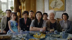 The character Billi, played by Awkwafina, sits at a dining table, surrounded by her family.